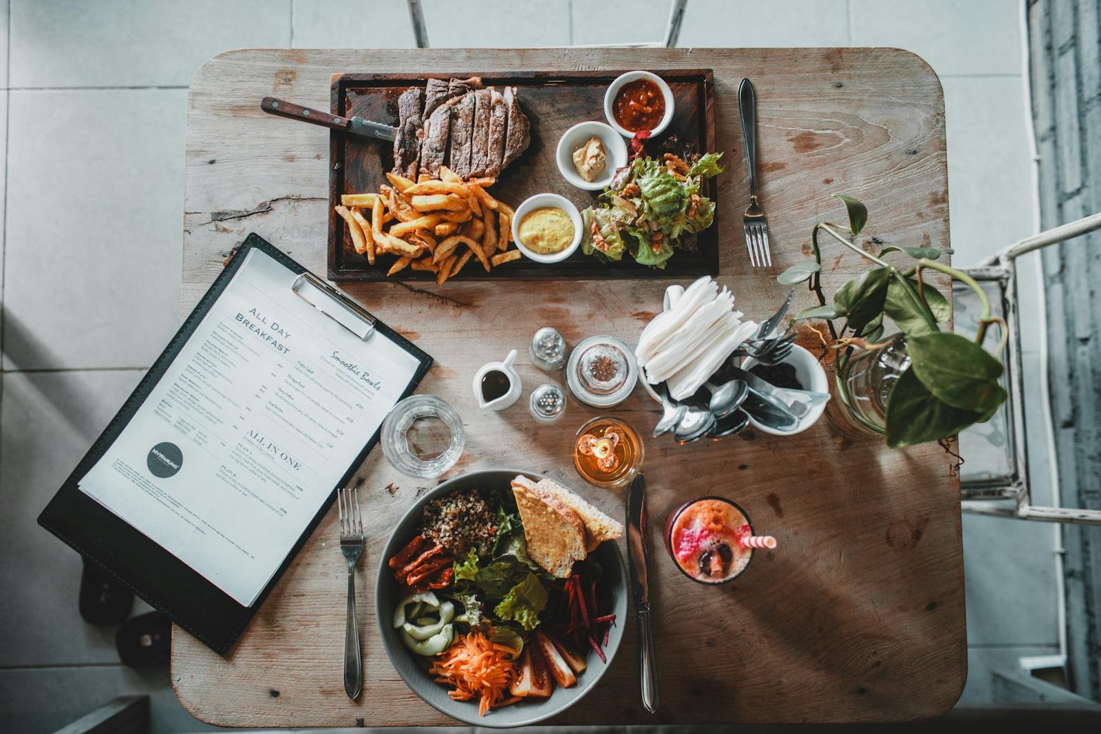 Top view of wooden table with salad bowl and fresh drink arranged with tray of appetizing steak and french fries near menu in cozy cafe